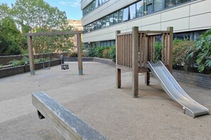 An outdoor playground with a slide and swings, surrounded by a paved area and greenery. - Wembley Park 1 Bed | Sleep 4 | Gym, Workspace (Greater London)