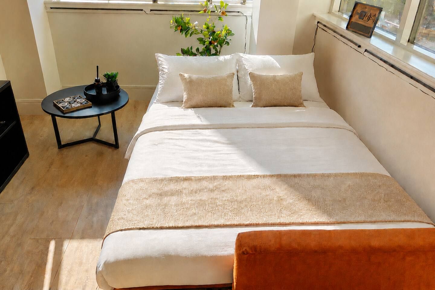 Ein ordentlich bezogenes Bett mit weißer Daunendecke und zwei Kissen, ein kleiner runder Tisch mit einer Pflanze und ein Fenster mit Blick ins Freie.