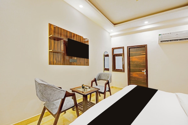 A hotel room with a bed, two chairs, a small table, a flat-screen TV mounted on a wooden panel, a door, and an air conditioning unit.
