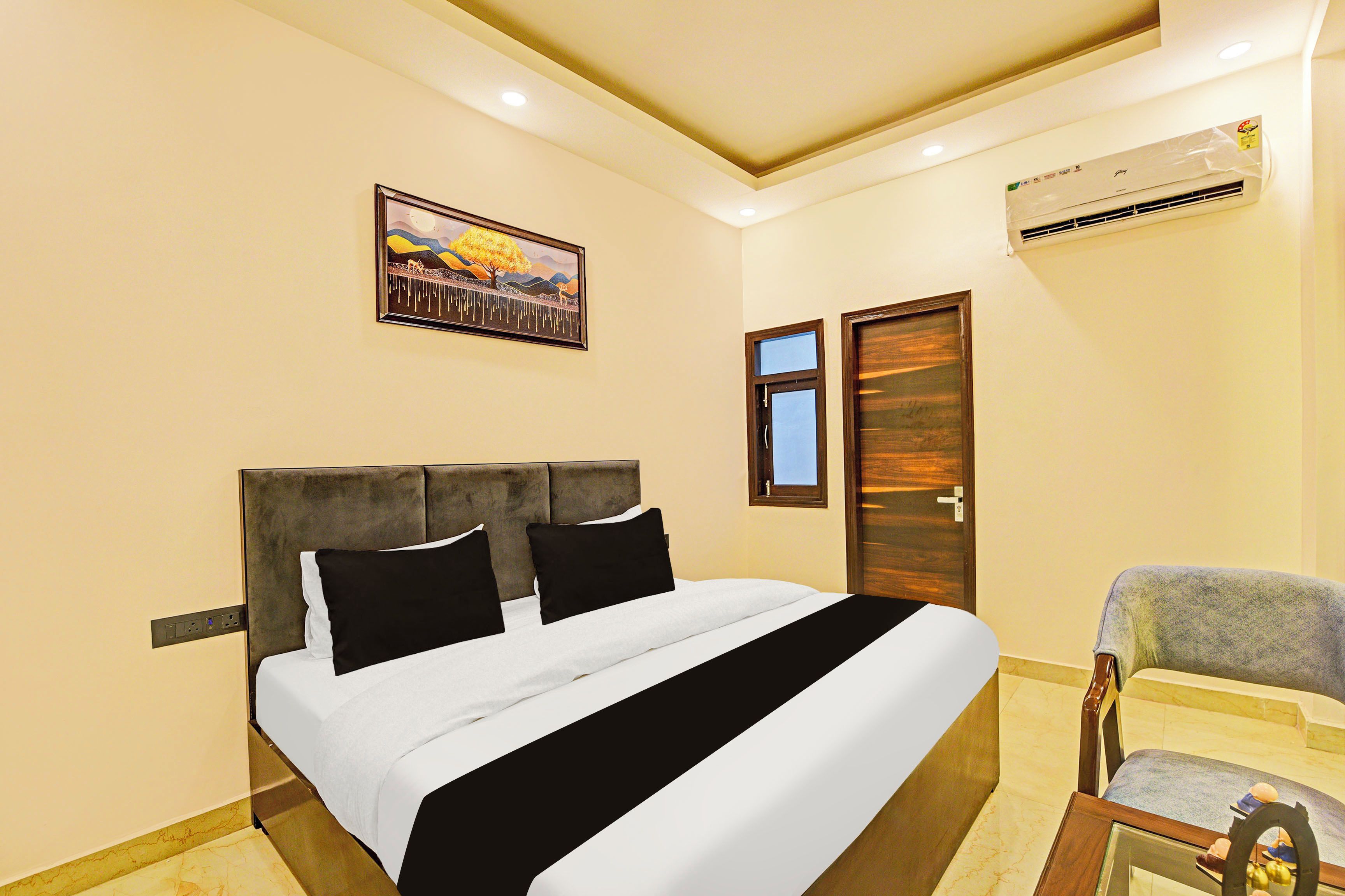 Habitación de hotel con una cama, una silla, un equipo de aire acondicionado, una puerta y un cuadro en la pared.