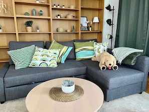 Ein graues Ecksofa mit gemusterten Kissen, ein runder Couchtisch mit gewebter Unterlage und ein kleiner Topfpflanze.