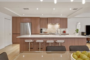 A modern kitchen with a stainless steel refrigerator, a marble countertop, and a built-in stove. There are four bar stools and a dining table with a bowl of lemons. - Luxurious beautifully renovated Coconut Grove retreat with private Hot tub. (Miami)