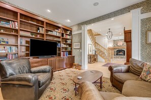 A spacious living room with a large flat-screen TV, leather sofas, a wooden bookshelf, and a staircase leading to an upper level. - Asheville Mountain Lodge by Biltmore; Creek, Hot Tub, Theater & Games (Asheville)