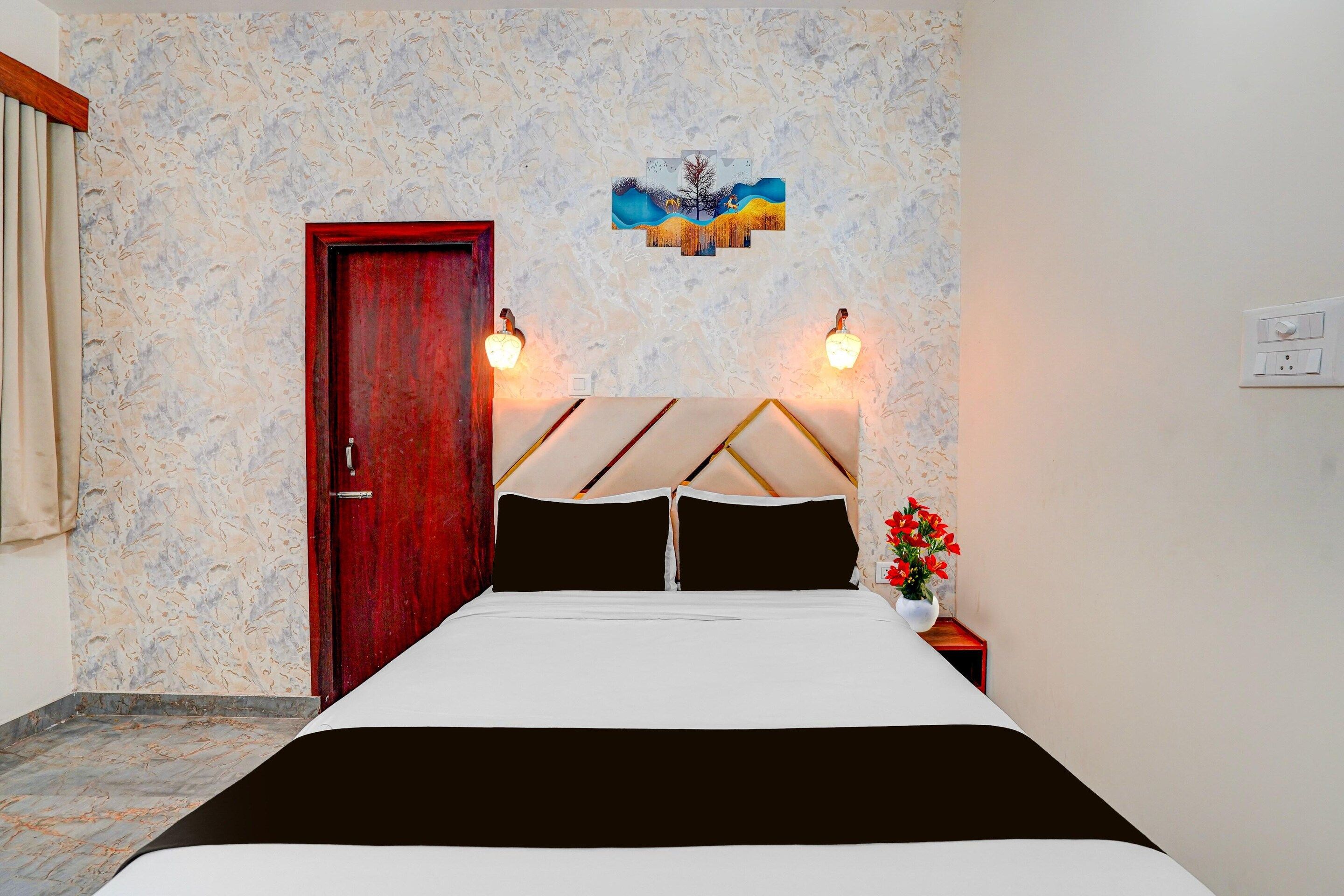 A bedroom with a bed, a wooden door, a wall-mounted light fixture, and a small table with a vase of flowers.