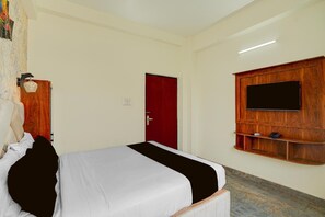 A hotel room with a bed, a red door, a mounted TV, and a wooden shelf.