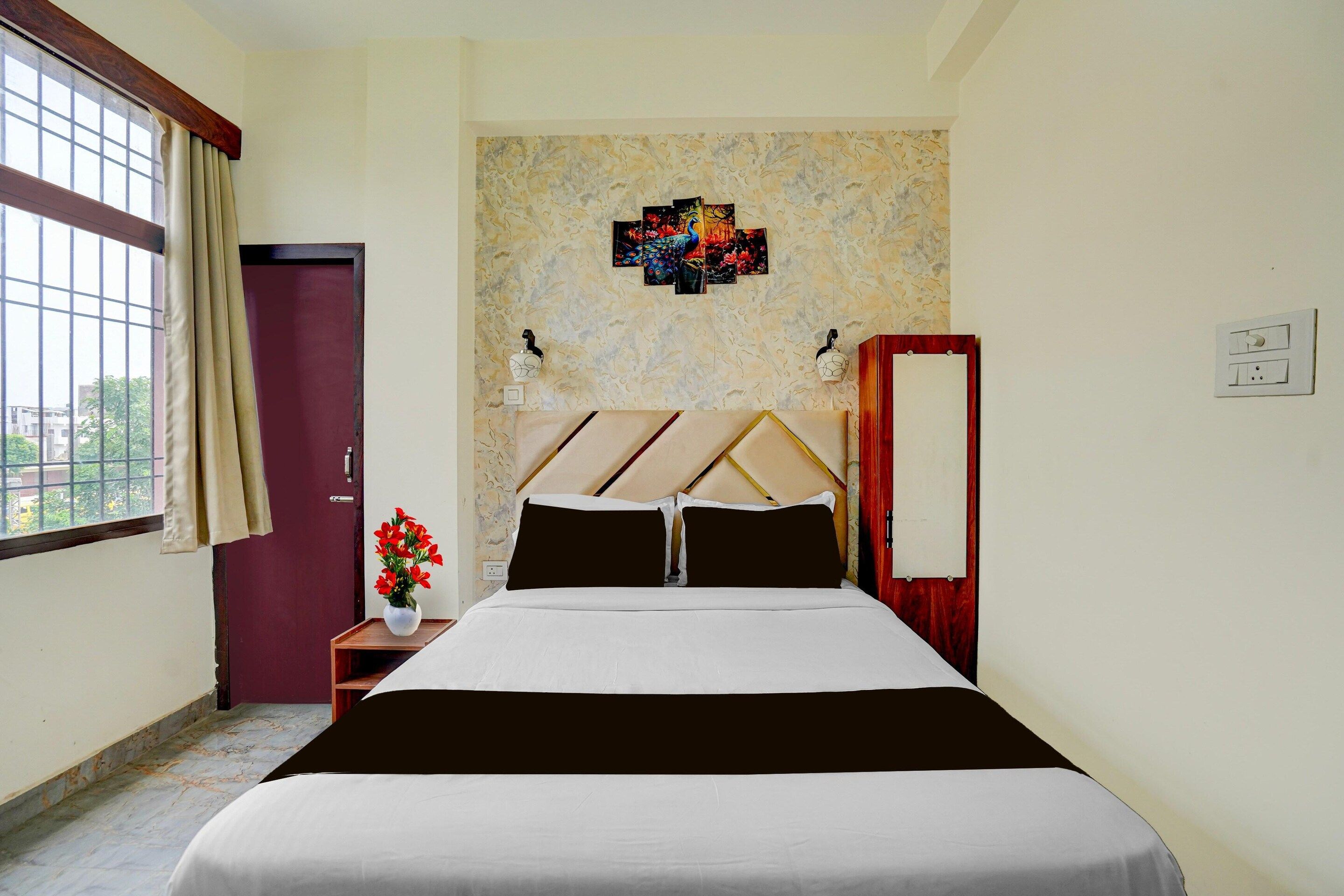 Una habitación de hotel con una cama, una mesita de noche con un florero y una ventana con cortinas, además de una pared con un adorno decorativo.