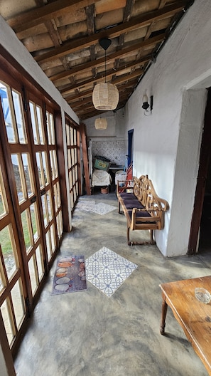 A room with a high ceiling, wooden beams, a woven ceiling light, a patterned floor mat, a wooden bench, and a table.