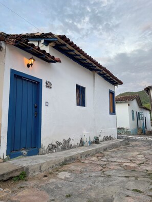 A white building with a blue door and window shutters, a stone-paved street, and a clear sky.