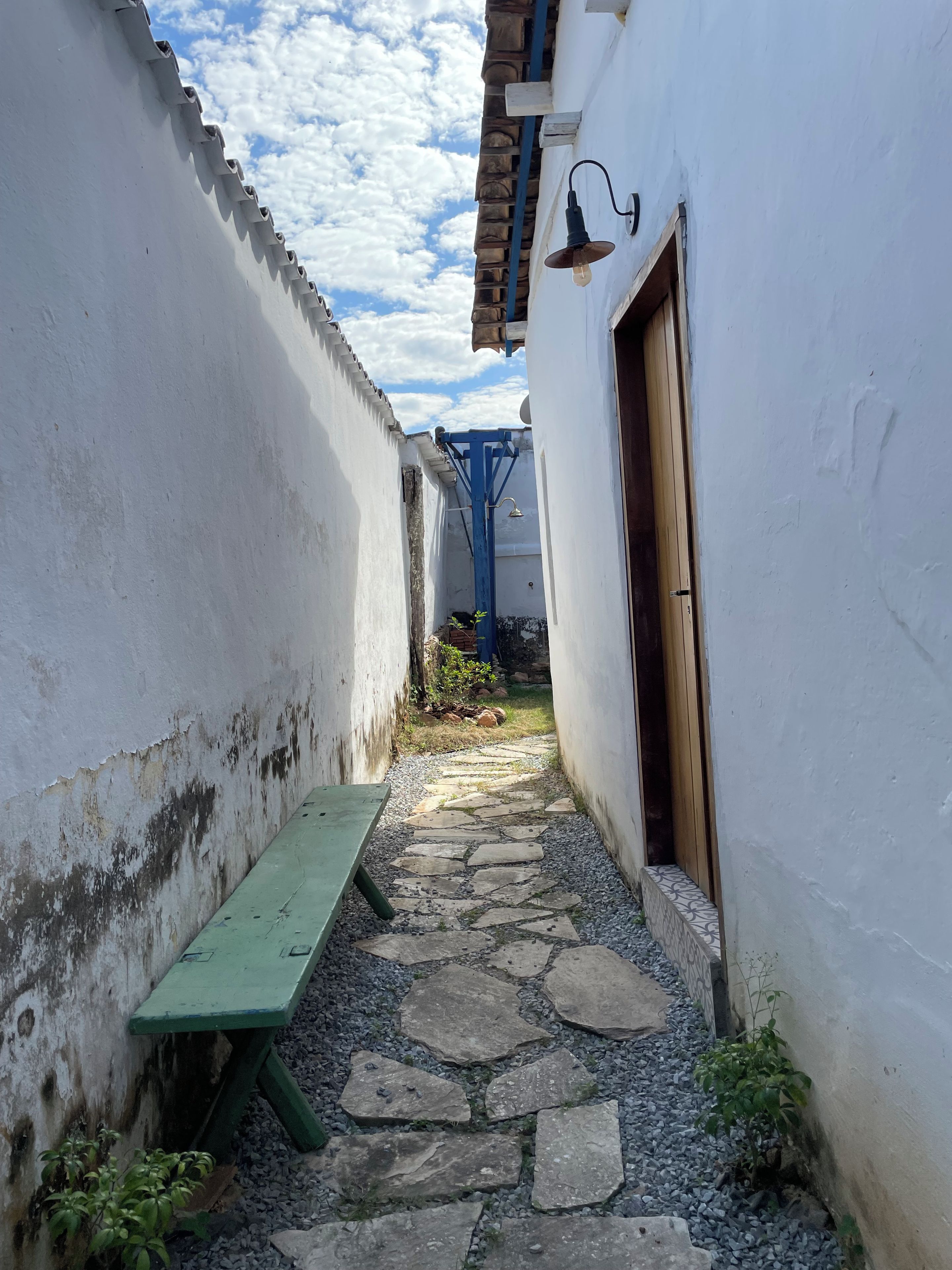 A narrow alleyway with a stone pathway, a green bench, and a building with a blue door and light fixture.