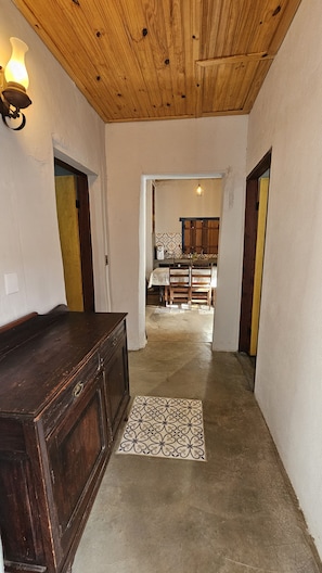 A hallway with a wooden ceiling, a dark wooden bench, and a patterned rug leading to a kitchen area with wooden cabinets and a table.