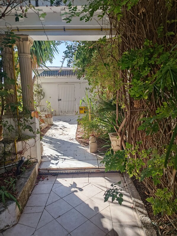 Un camino conduce a la entrada de un edificio con suelo de baldosas, rodeado de vegetación y un techo tradicional de azulejos.