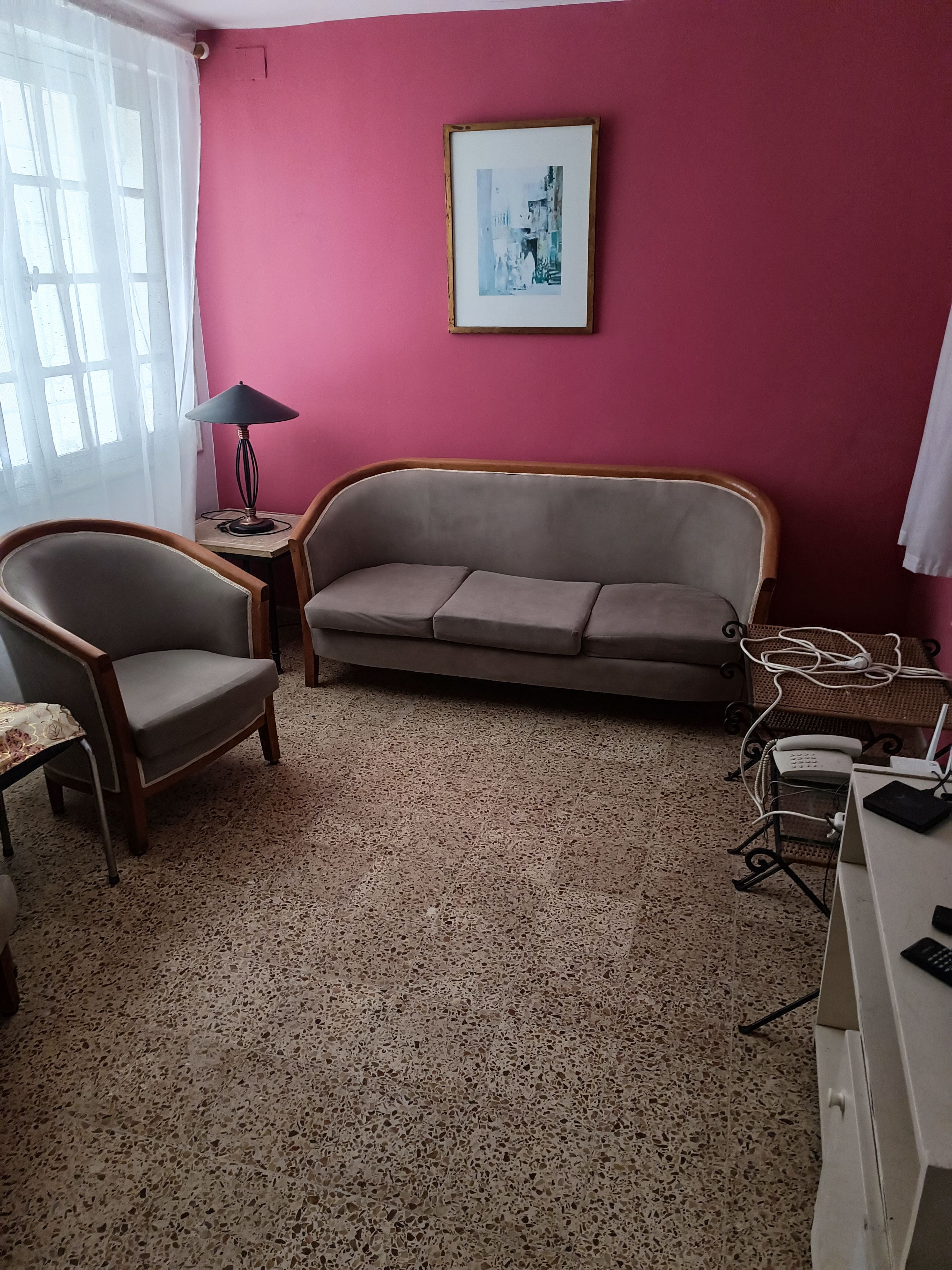 A room with a sofa, a chair, a table with a lamp, a framed picture on the wall, and a desk with a phone and computer.