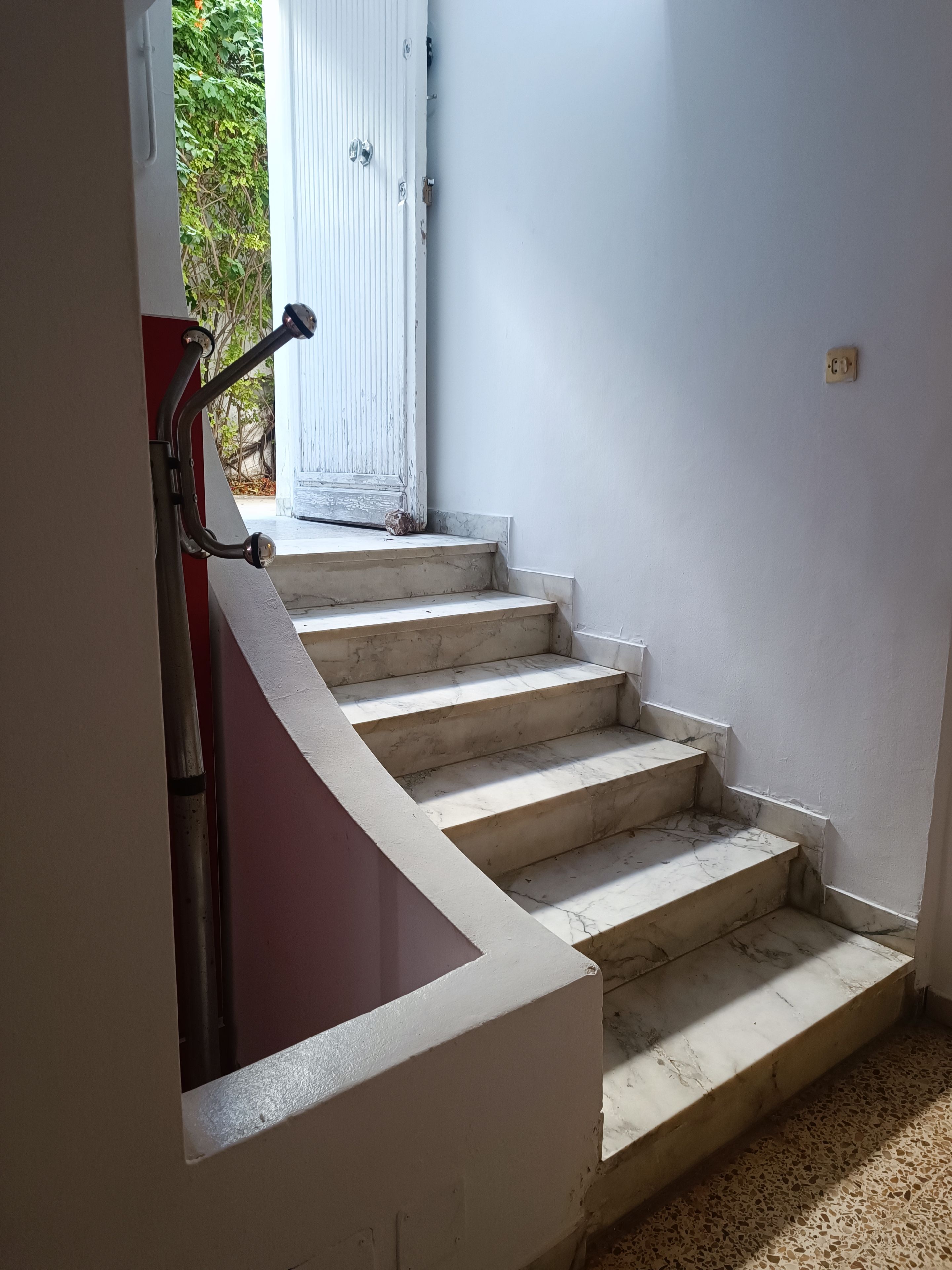 Un escalier aux murs blancs et à la rampe rouge, menant à une porte.