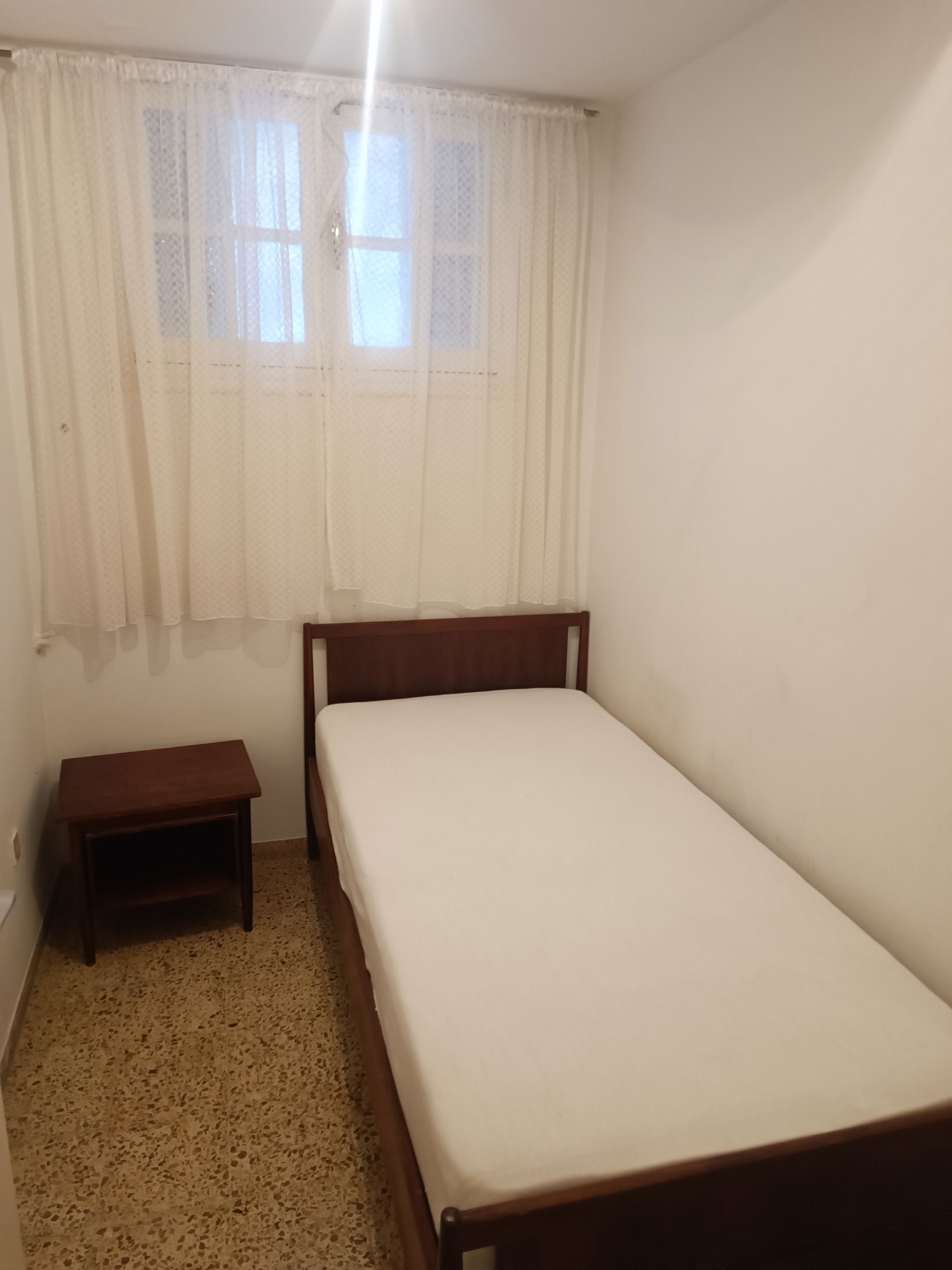 Una habitación pequeña con cama individual, cabecera y mesita de noche de madera, ventana con cortinas traslúcidas y suelo moteado.