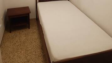 Una habitación pequeña con cama individual, cabecera y mesita de noche de madera, ventana con cortinas traslúcidas y suelo moteado.