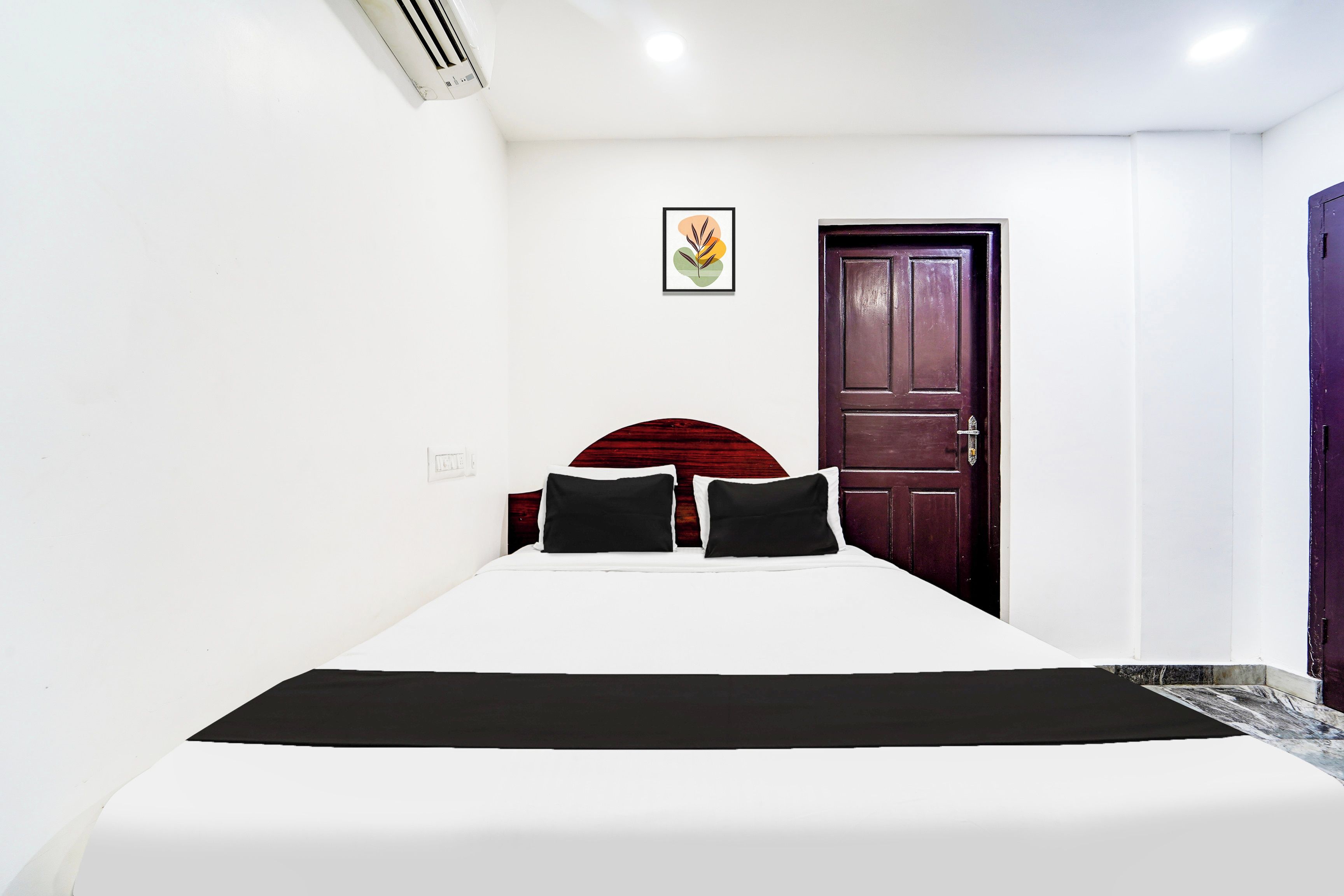 Habitación de hotel con una cama individual, cabecera de madera oscura, pared blanca, un cuadro enmarcado y una puerta cerrada.