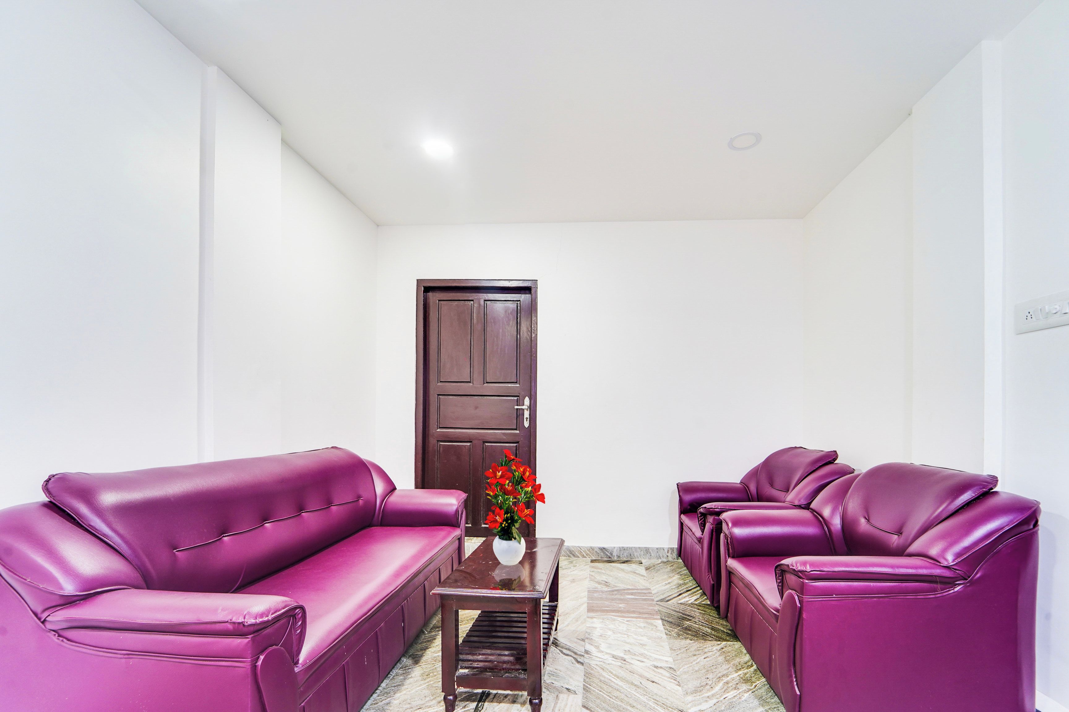 A room with a dark wooden door, a small table with a vase of flowers, and purple leather sofas.