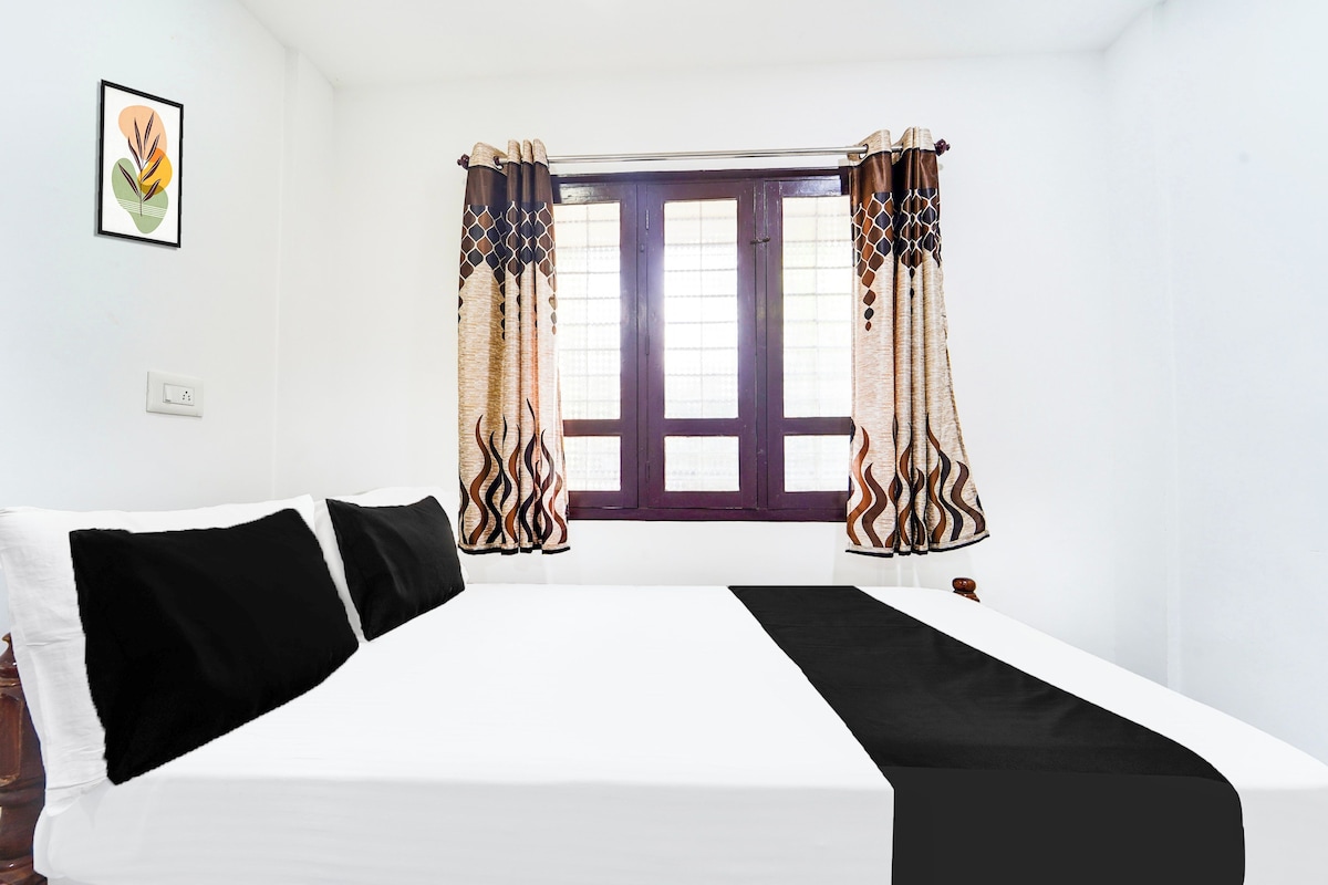 A room with a bed, a window with curtains, a wall-mounted light switch, and a framed picture.