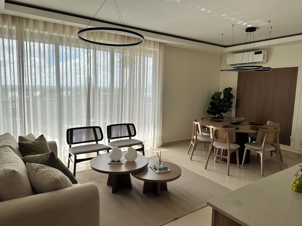 A modern living room with a sofa, two coffee tables, and a dining area with chairs and a table. - New Designer 3BR • Mountain Views • Near STI (Santiago de los Caballeros)
