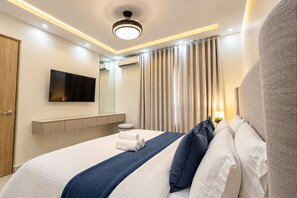A modern hotel room with a large bed, a flat-screen TV, a mirror, a bedside table, and a window with curtains. - New Designer 3BR • Mountain Views • Near STI (Santiago de los Caballeros)