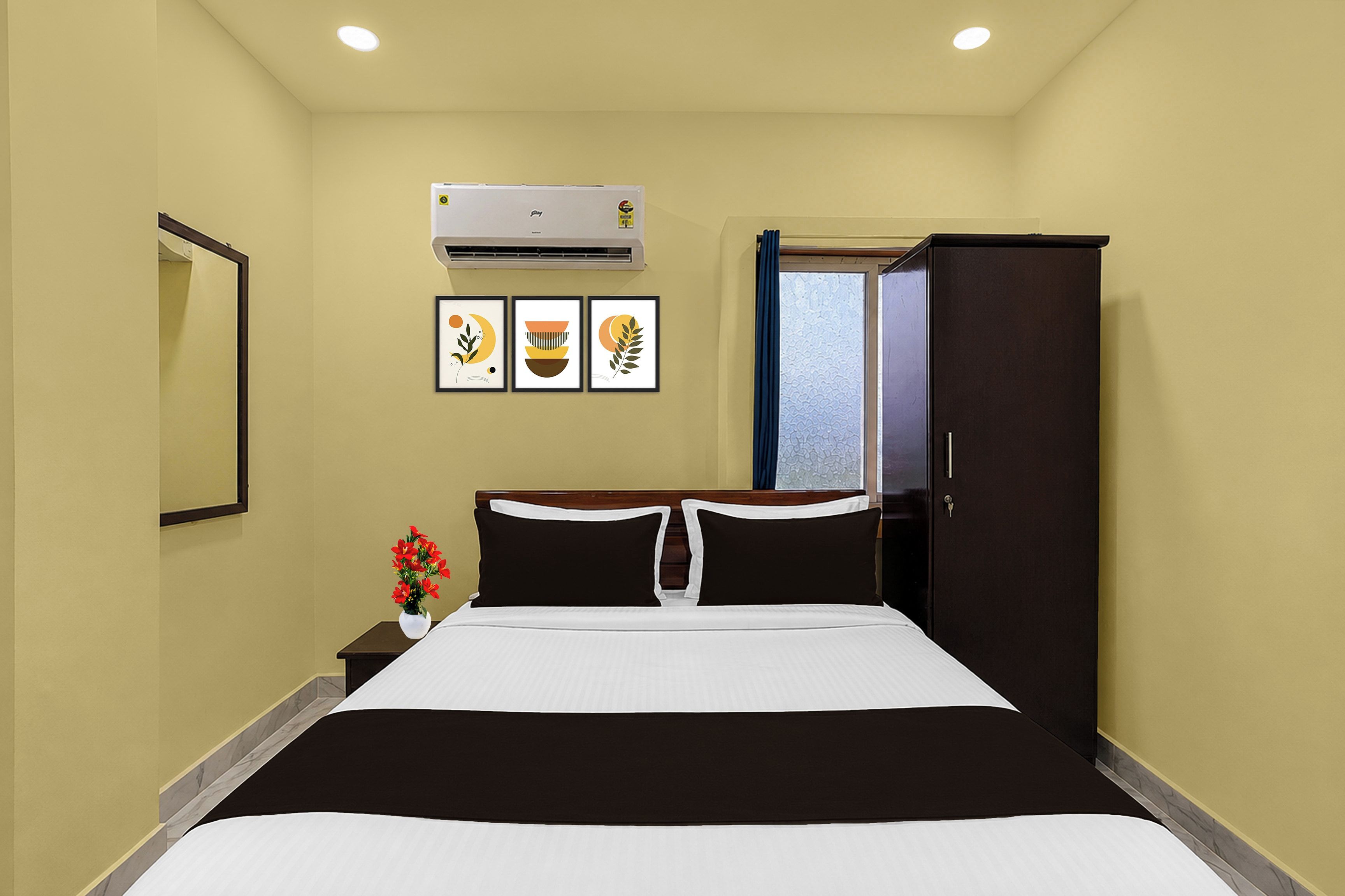 A hotel room with a bed, a wardrobe, an air conditioning unit, and three framed artworks on the wall.