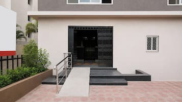 Una entrada de edificio moderno con una escalera que lleva a una puerta con portón, un pequeño jardín con plantas y pavimento de baldosas.
