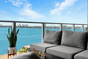 A balcony with a grey sofa, potted plants, and a view of a city skyline across a body of water. - Azure Crown Apartment (Miami)