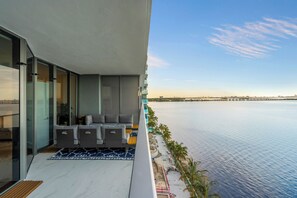 A balcony with a sofa and chairs, overlooking a calm body of water and a distant city skyline. - Azure Crown Apartment (Miami)
