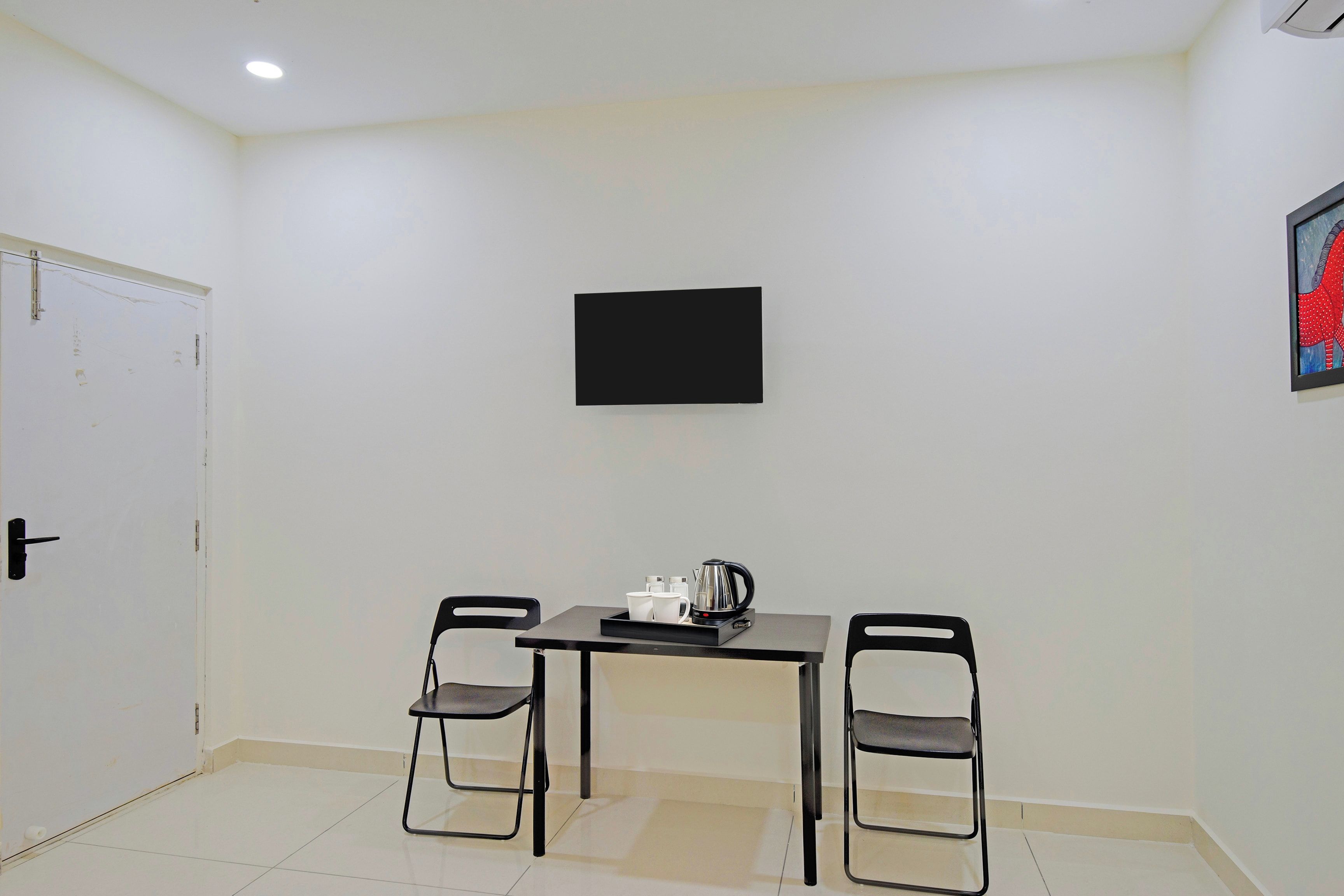 A room with a white door, a black-framed TV on the wall, a table with two chairs, and a kettle on the table.