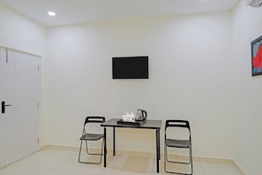 Una habitación con una puerta blanca, un televisor con marco negro en la pared, una mesa con dos sillas y un hervidor sobre la mesa.
