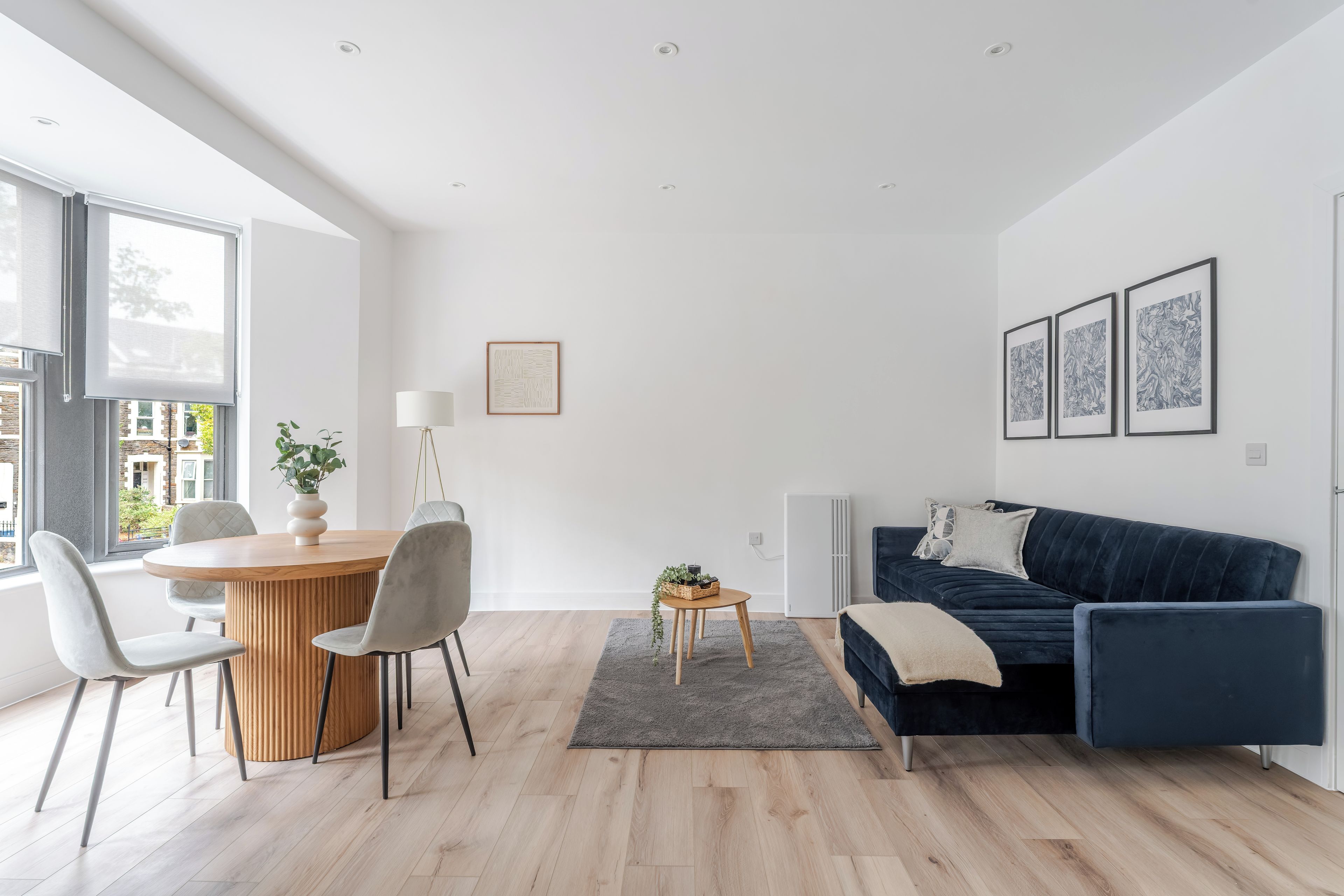 A modern living room with a dark blue sofa, a round wooden dining table with chairs, a grey rug, and a white radiator.