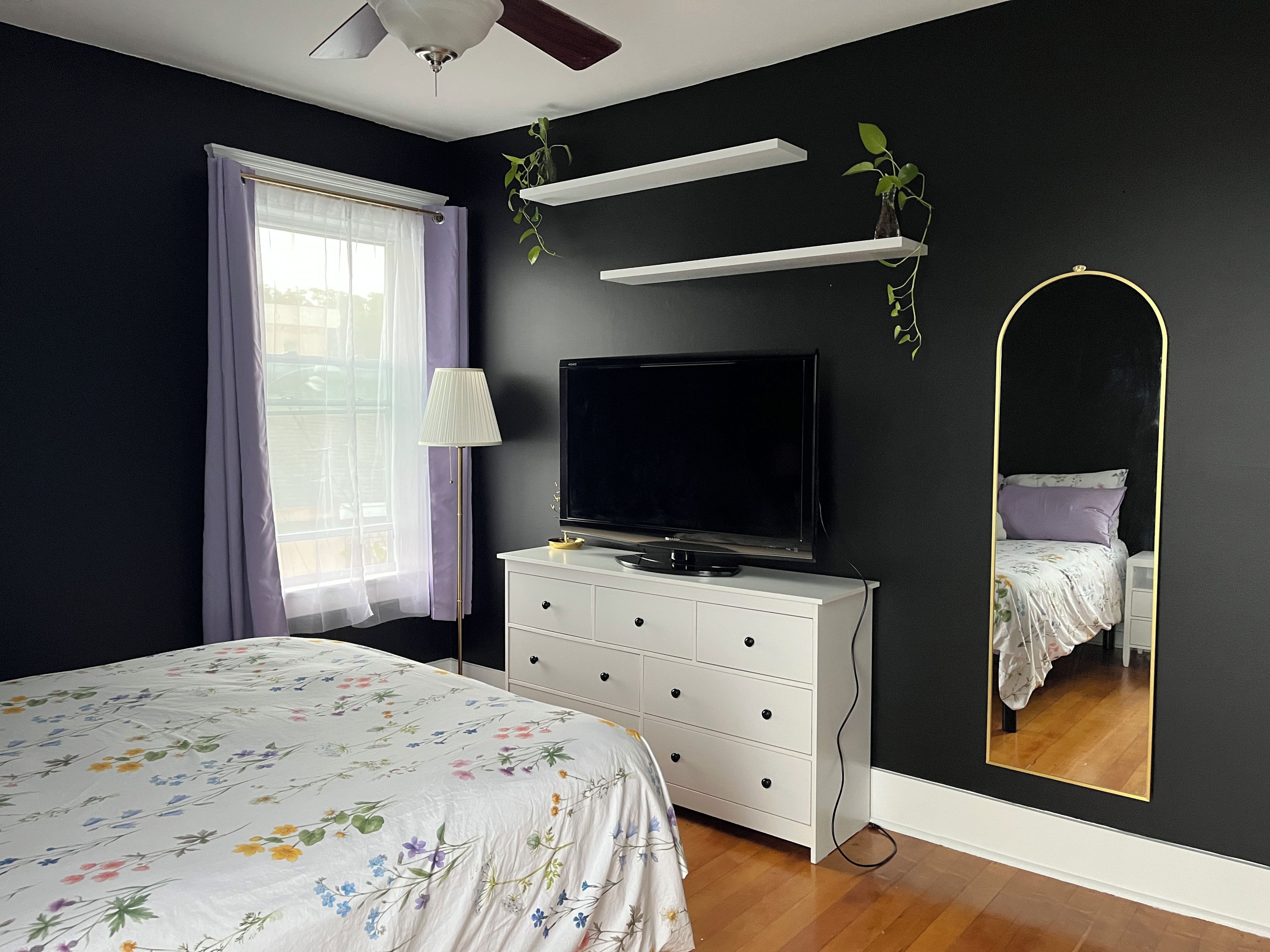 Une chambre à coucher avec un lit, une télévision, une commode, un miroir et une fenêtre avec des rideaux.