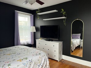 Un dormitorio con cama, televisión, cómoda, espejo y ventana con cortinas.
