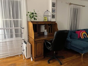 Una habitación con un escritorio de madera, una silla de oficina negra, un sofá azul con un cojín multicolor y un radiador blanco, además de una ventana con cortinas translúcidas.