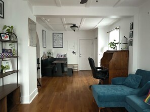 Una sala de estar con sofá azul, suelo de madera, una zona de comedor con mesa y sillas, y una estantería con cuadros enmarcados.