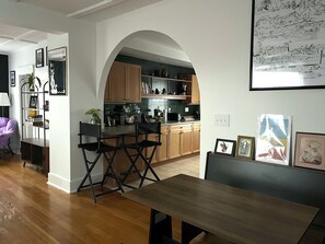 A modern kitchen with wooden cabinets, a dining area with a table and chairs, and a living room with a purple chair and a bookshelf.