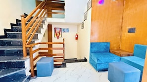 A hotel lobby with a wooden staircase, blue seating area, and a fire extinguisher on the wall. - Hotel Gurukripa Palace (Maheshwar)