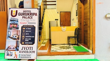 A hotel entrance with a reception desk, a signboard detailing room amenities, and a contact number for the hotel.