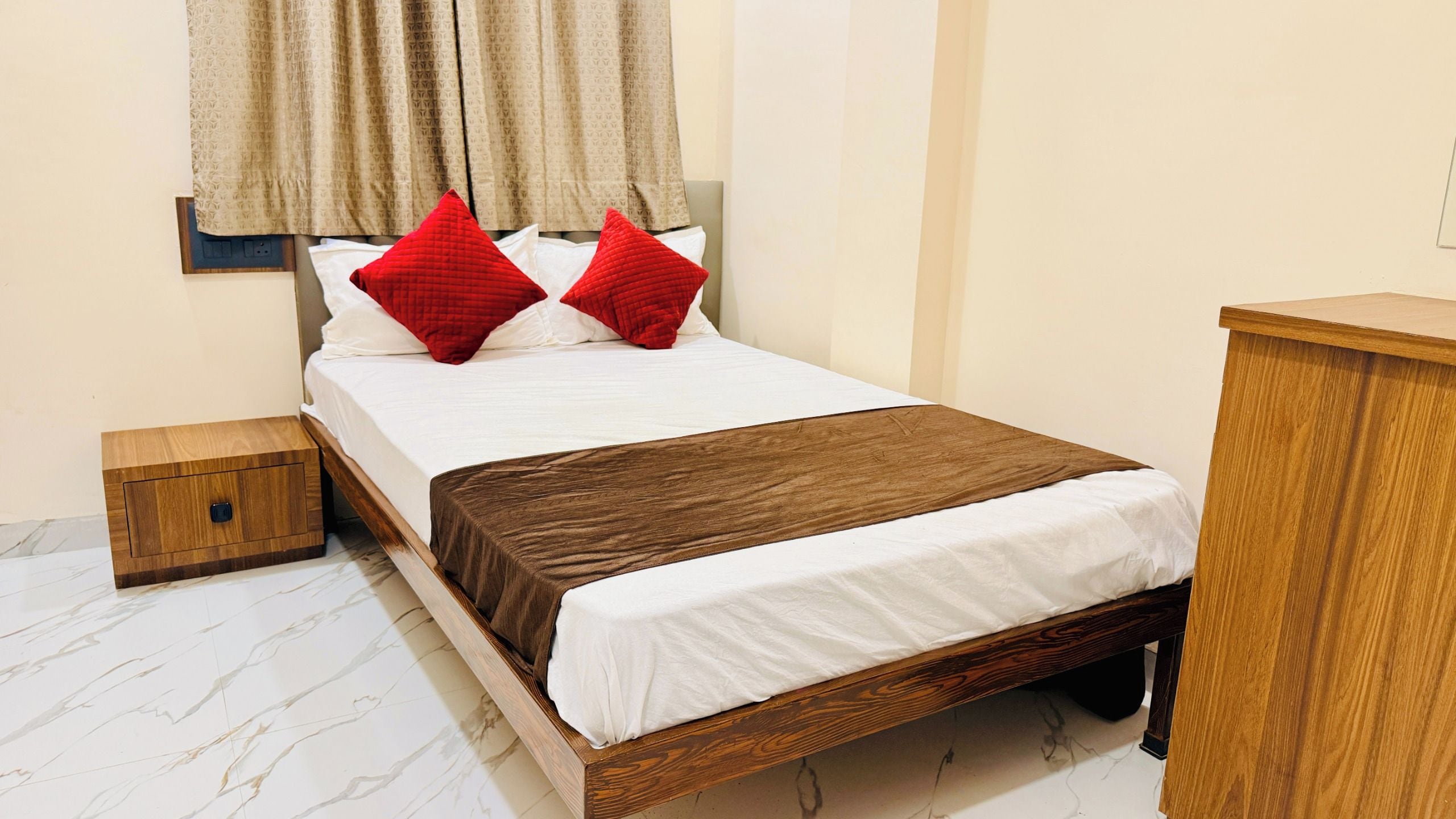 A neatly made bed with a wooden frame, white bedding, and two red pillows. There is a small wooden nightstand beside the bed, and a wooden dresser to the right.