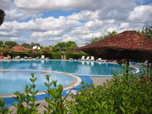 A swimming pool with lounge chairs and umbrellas, surrounded by greenery and a clear sky.