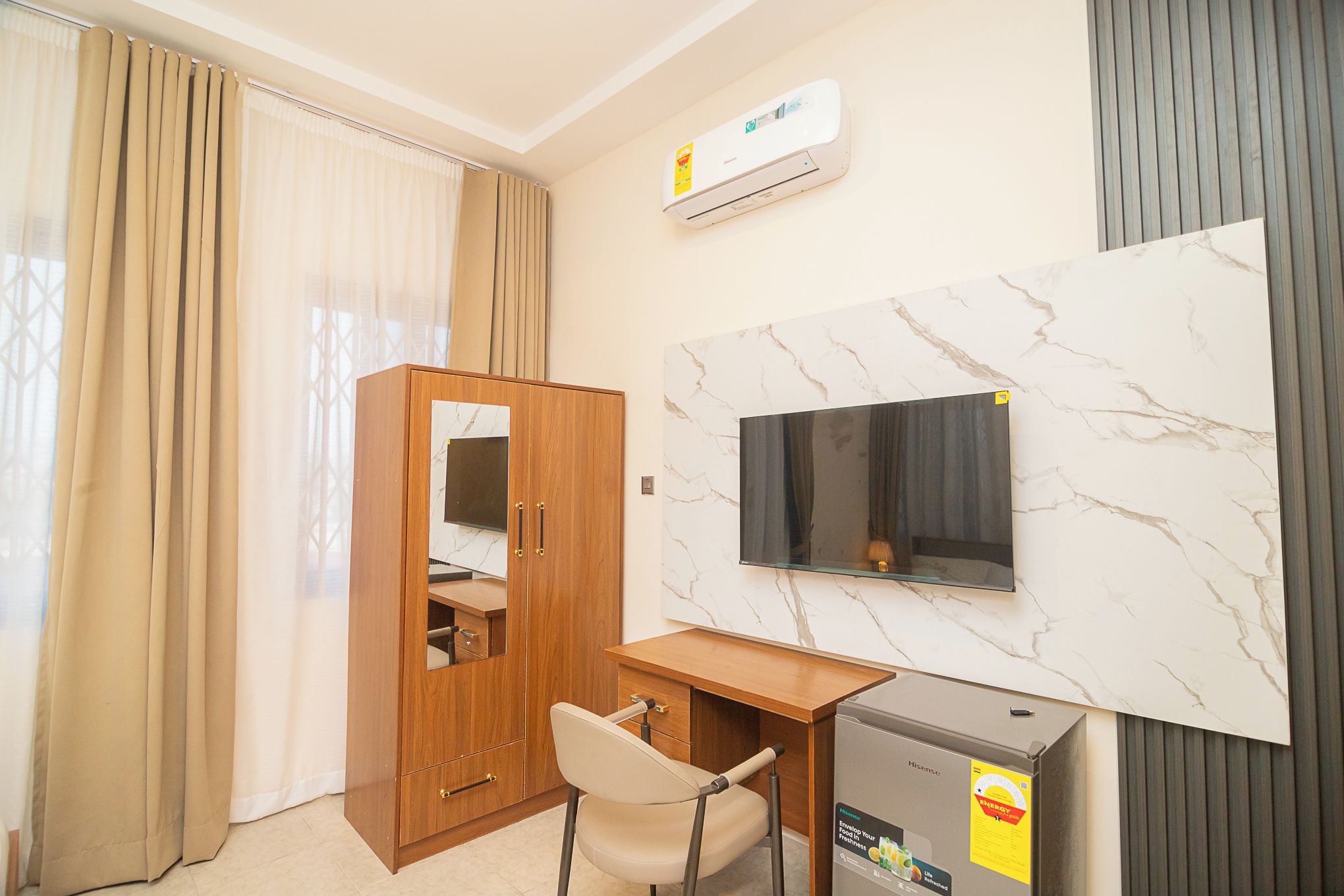 Habitación de hotel con armario de madera, un escritorio con silla, un televisor de pantalla plana montado en una pared de mármol y un refrigerador pequeño.