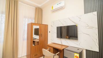 Habitación de hotel con armario de madera, un escritorio con silla, un televisor de pantalla plana montado en una pared de mármol y un refrigerador pequeño.