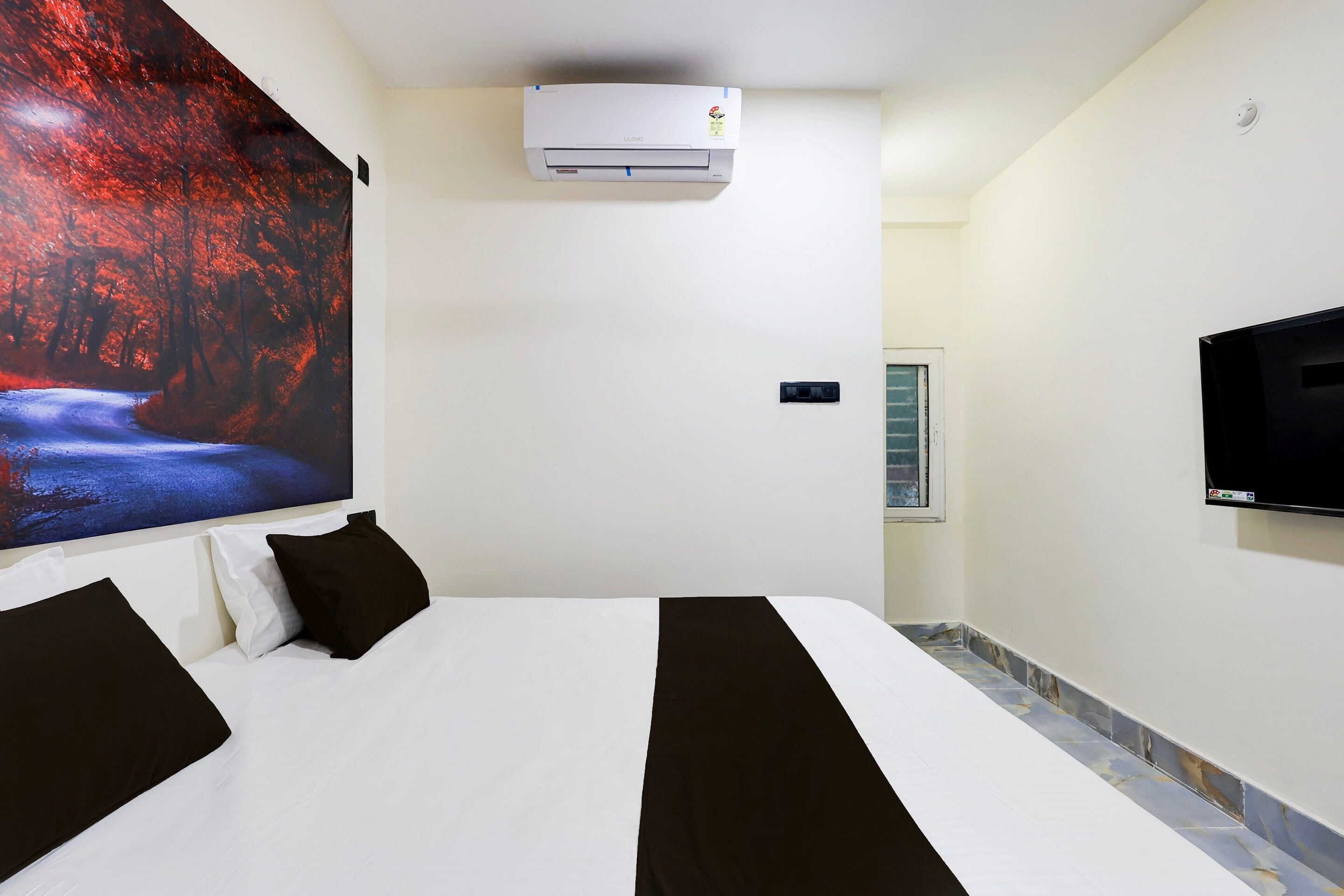 A hotel room with a bed, a mounted TV, an air conditioning unit, and a large painting on the wall.