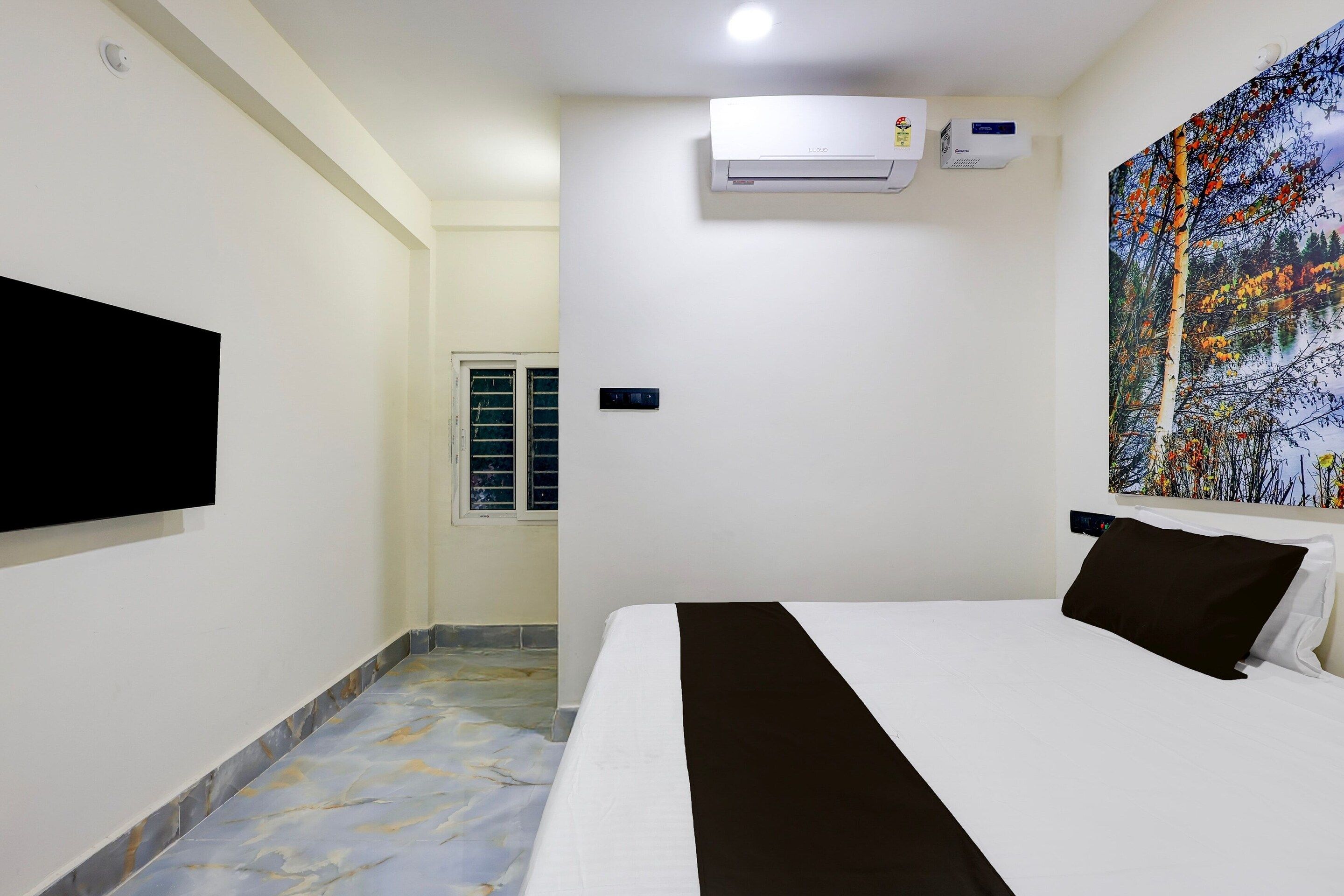 A hotel room with a bed, a mounted TV, an air conditioning unit, and a painting on the wall.