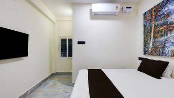 A hotel room with a bed, a mounted TV, an air conditioning unit, and a painting on the wall.