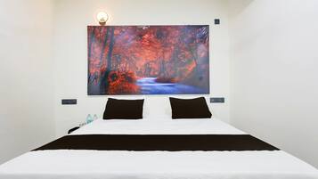 A hotel room with a large bed, two black pillows, a nightstand with a lamp, and a wall-mounted painting of a forest scene.