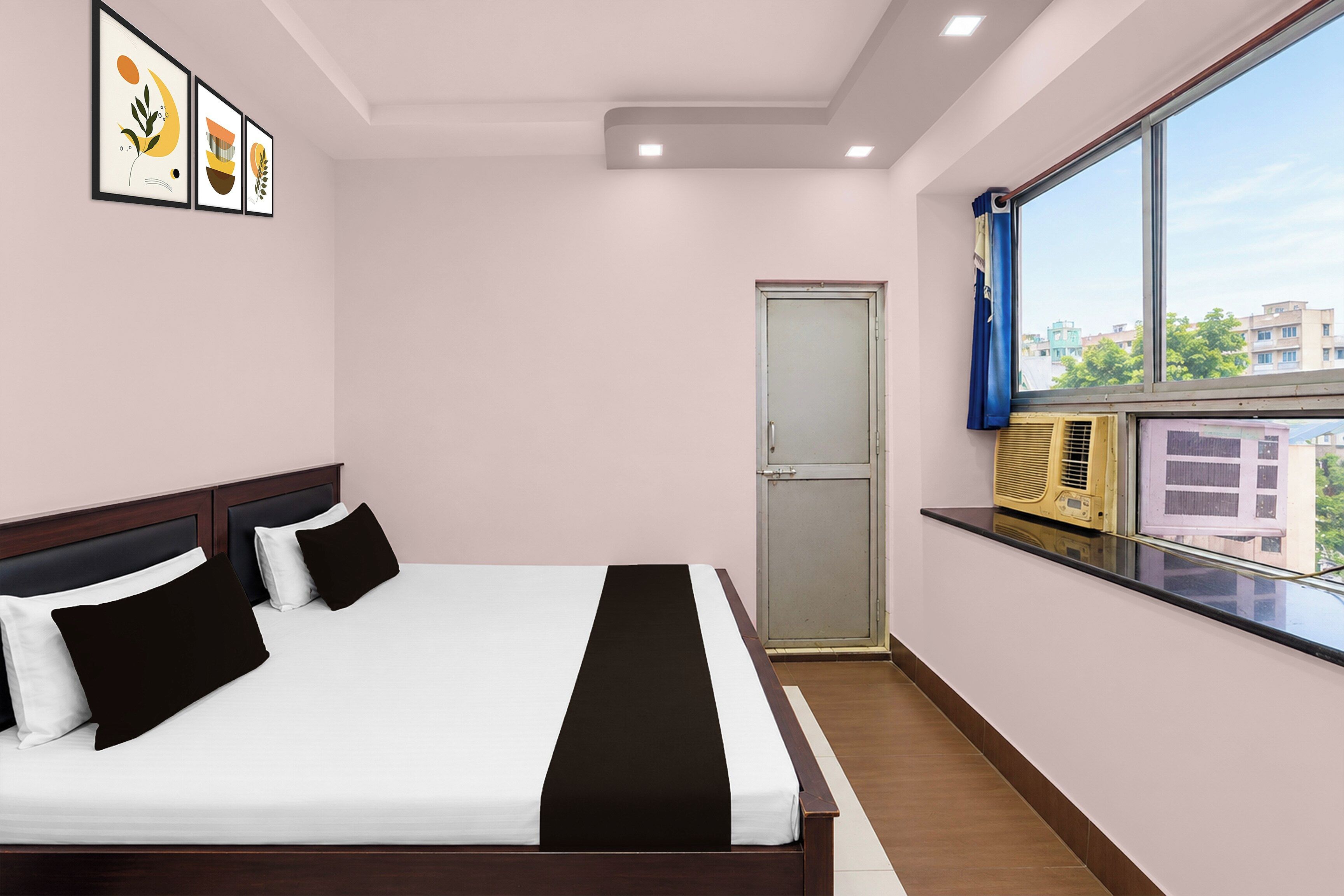 A hotel room with a bed, a window with a view, a door, and framed artwork on the wall.