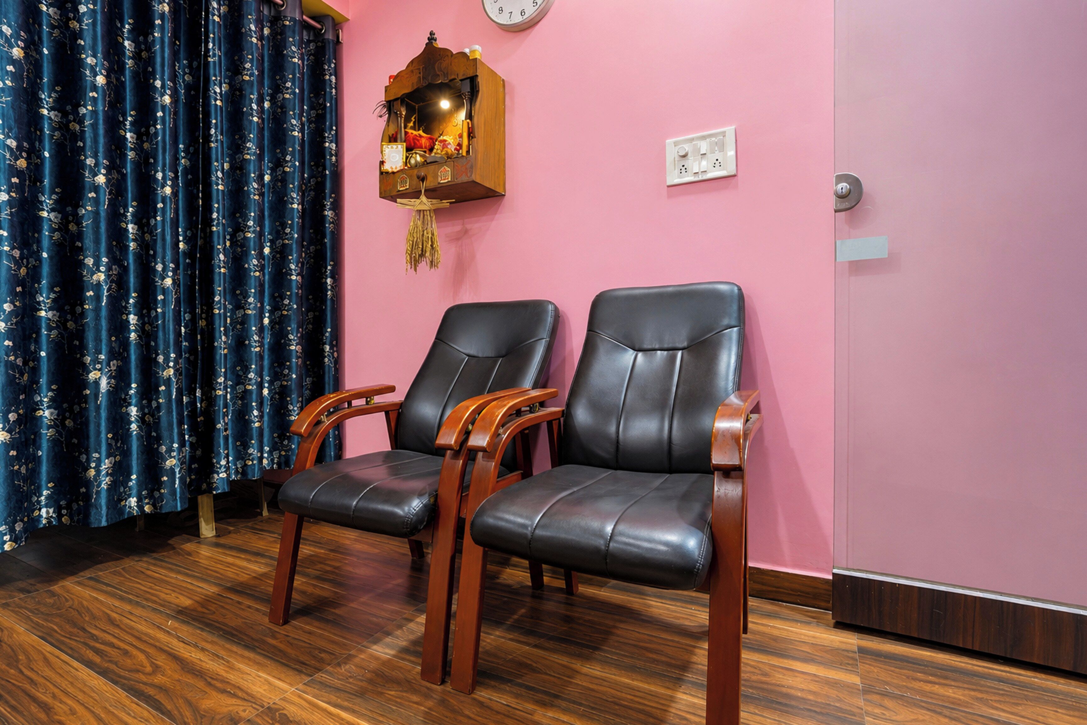 A room with two leather chairs, a wooden shelf with decorative items, a clock on the wall, and a power outlet.