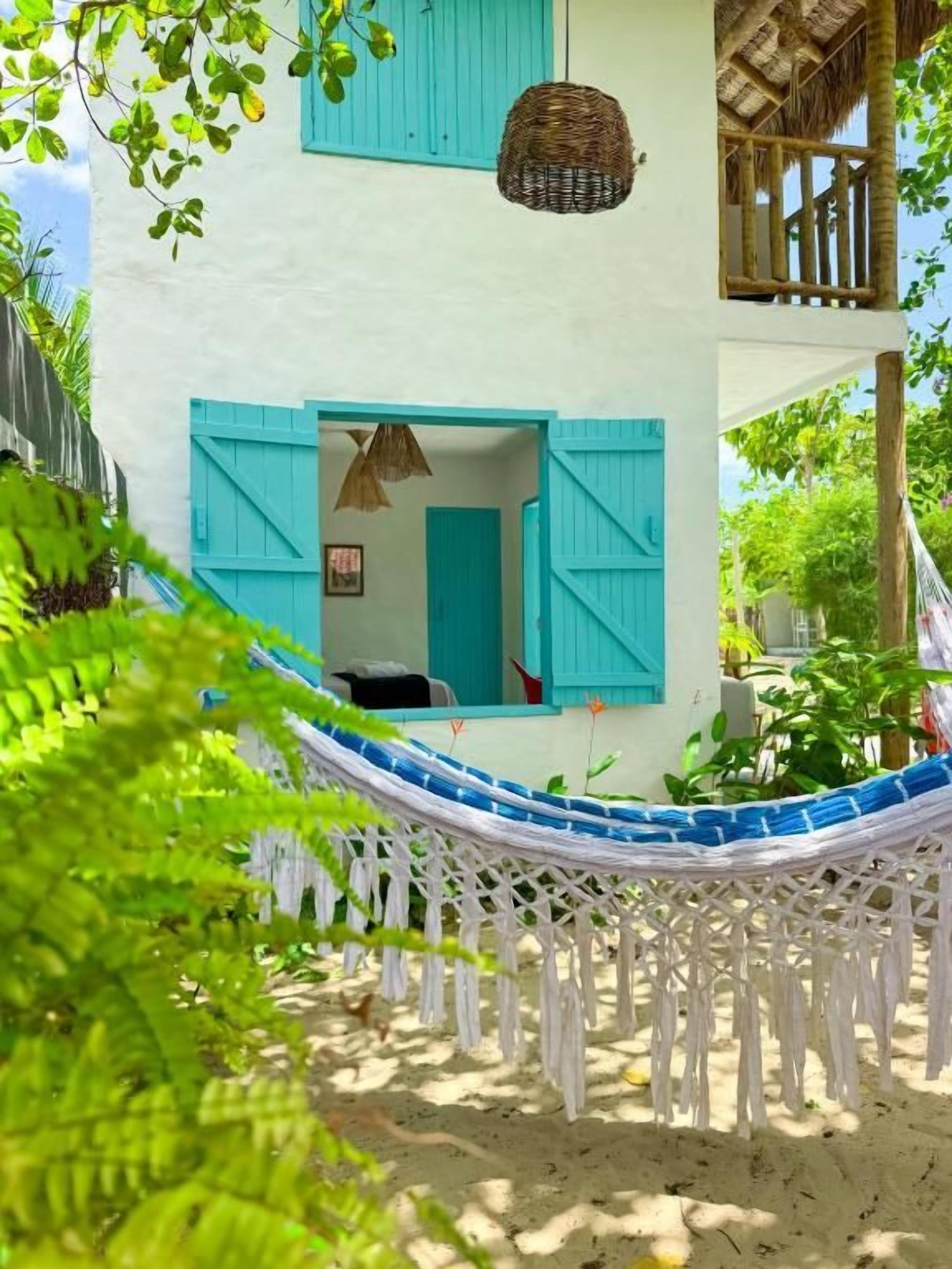 Uma rede esticada entre dois edifícios, sendo um deles branco com persianas azuis, e uma estrutura com telhado de palha.