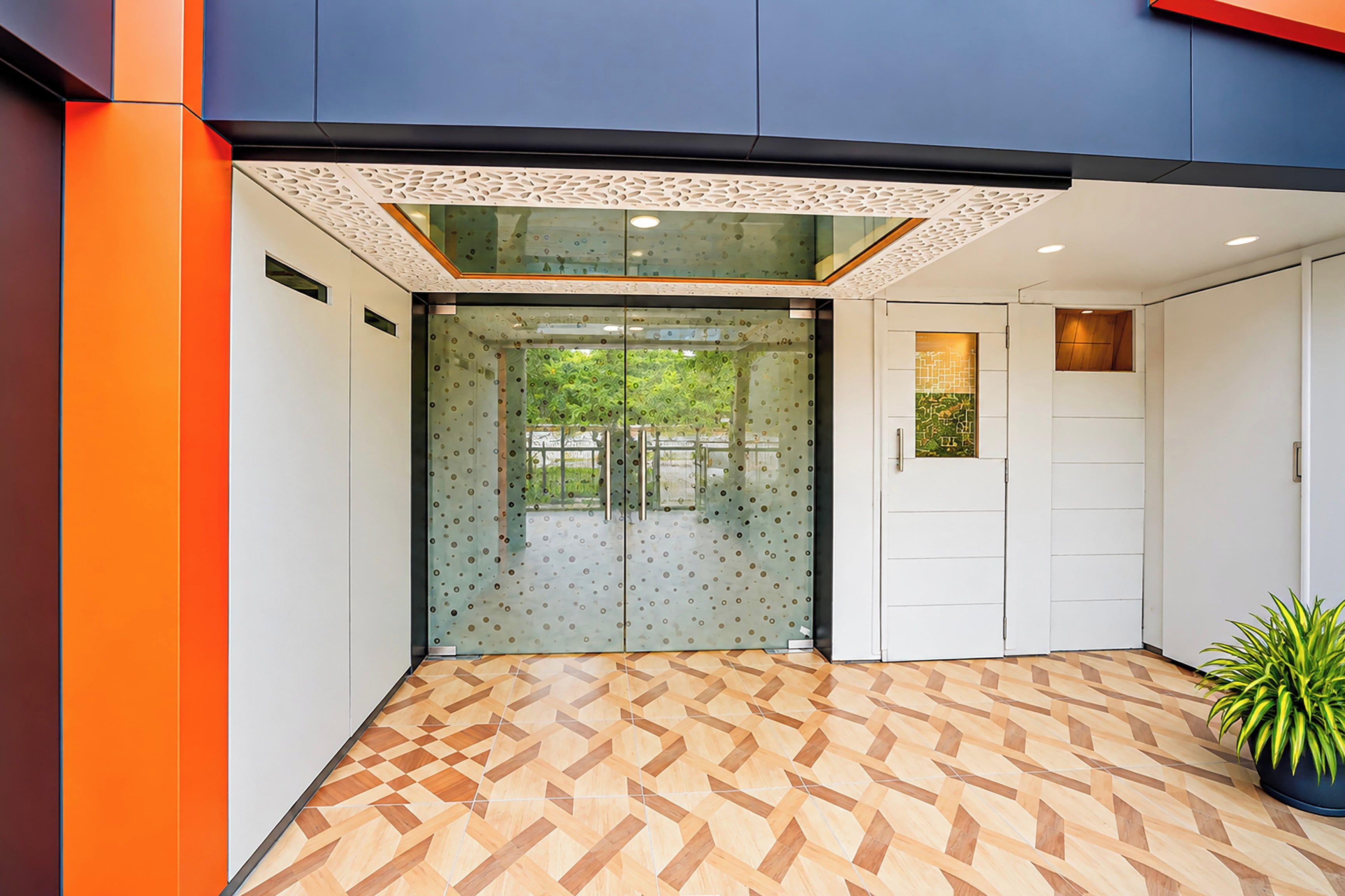 Entrada de um edifício moderno com piso de padrão geométrico, porta de vidro com textura fosca e um vaso de planta à direita.